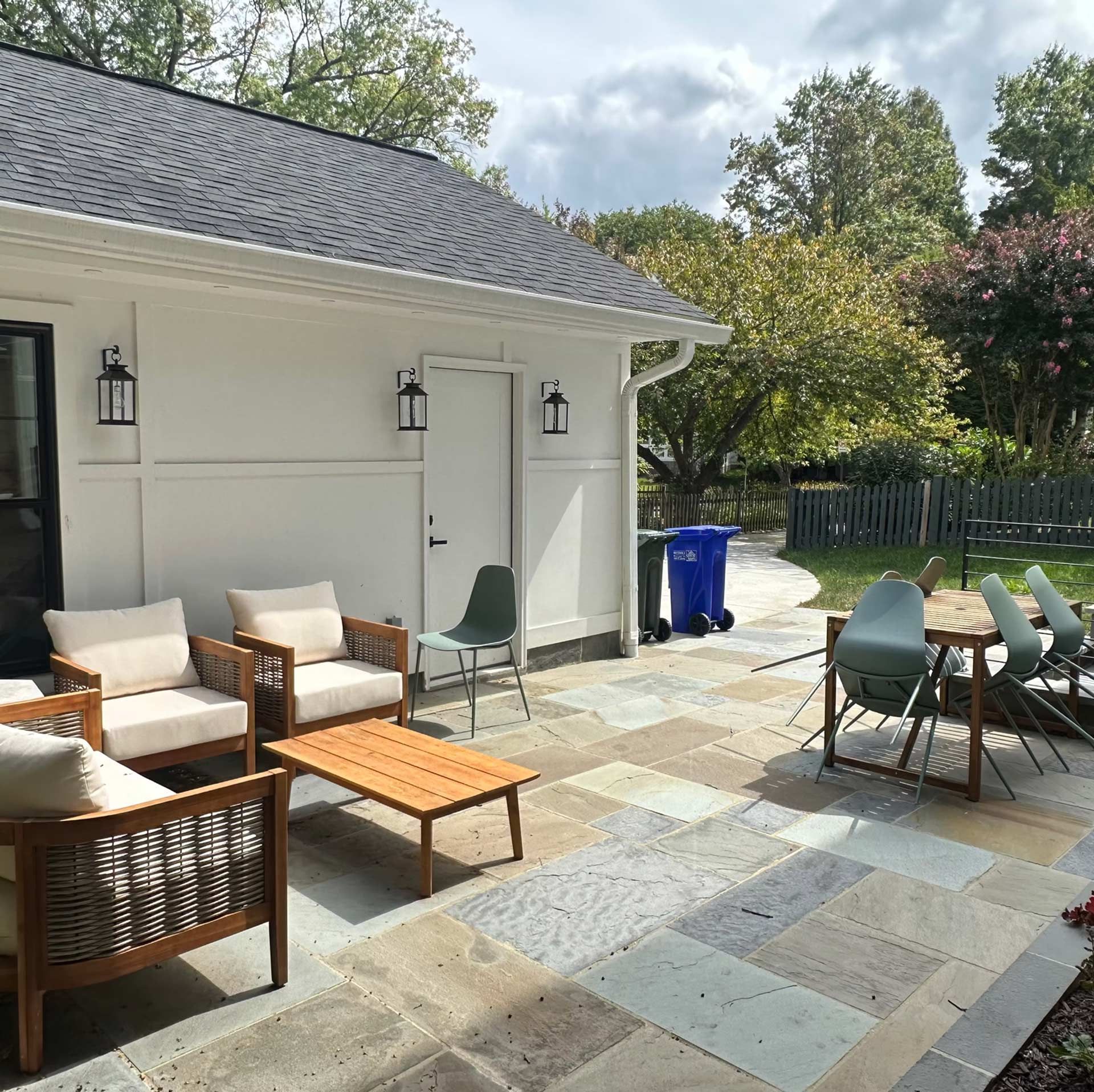 A stone patio with outdoor seating, a wood coffee table, and a dining set beside a white house with black light fixtures.