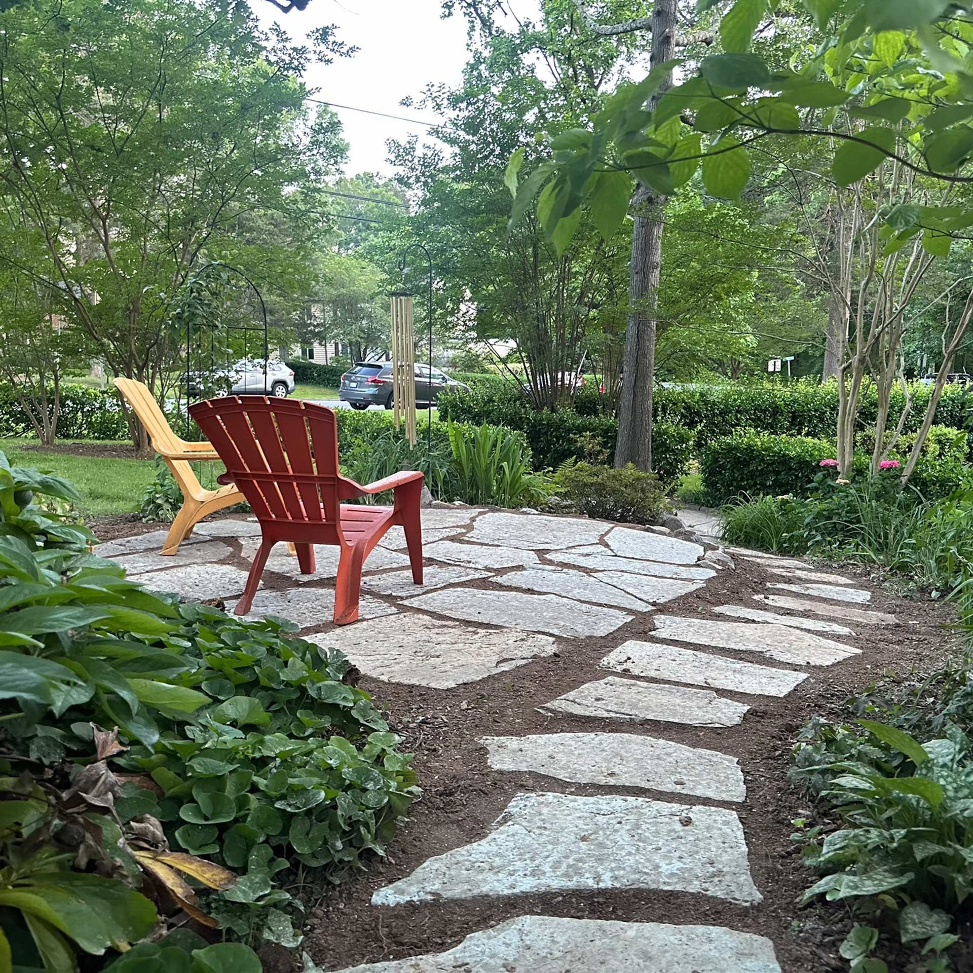 A stone path leads to a small patio with a red and a yellow Adirondack chair in a lush, green garden.