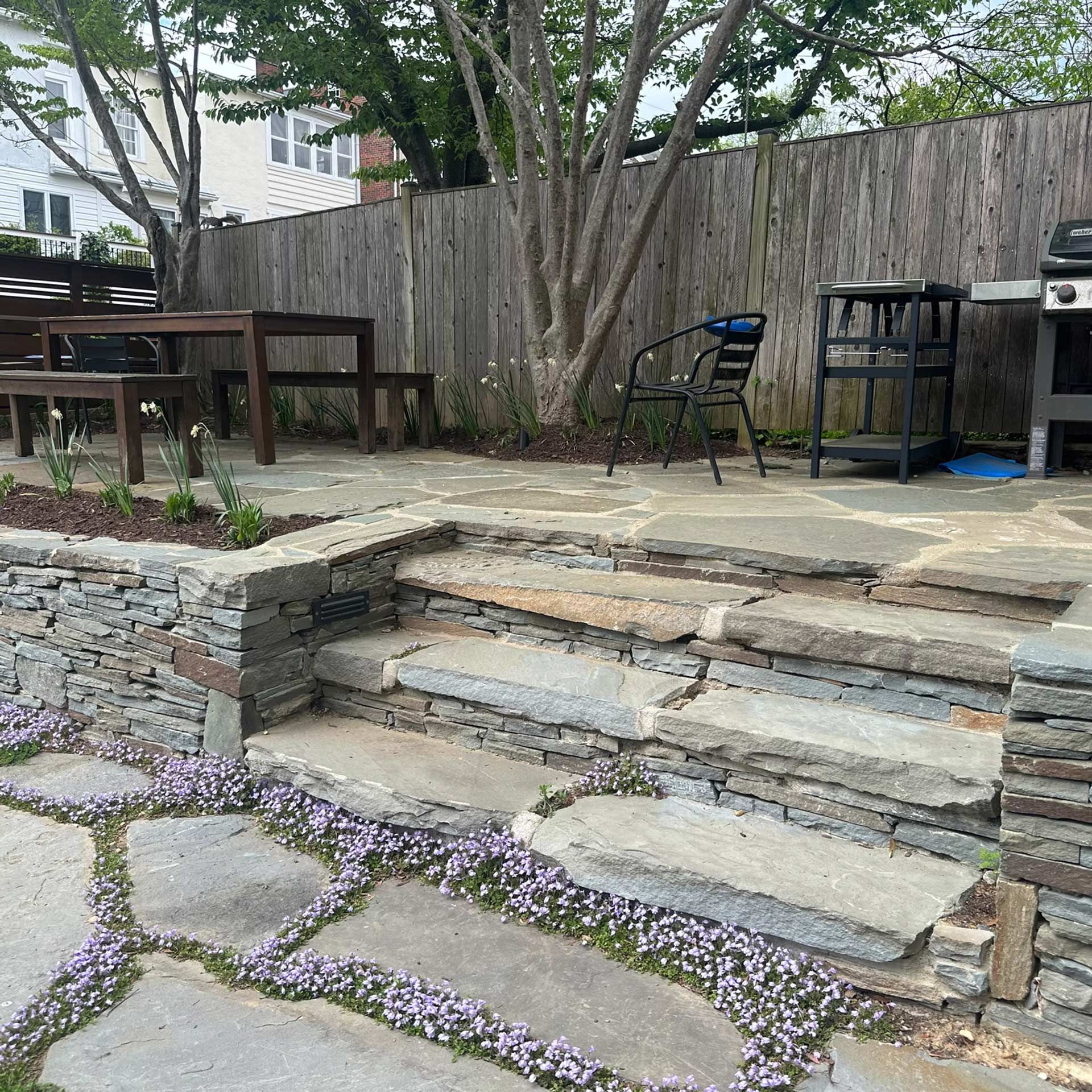 A flagstone patio features a set of stone steps leading to an upper level with a dining table, a grill, and a fence.
