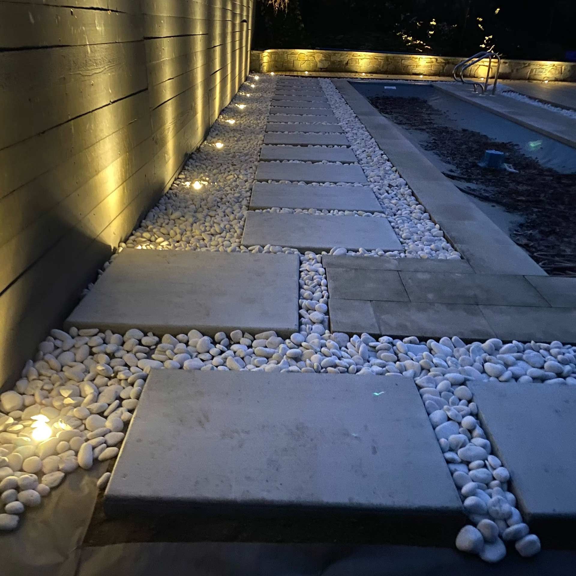 A walkway with rectangular concrete pavers and white stones, illuminated by landscape lights at night.