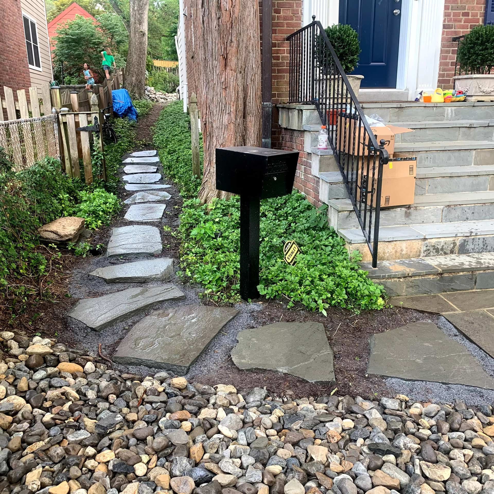 A stone pathway winds along a brick house next to a large tree and a black mailbox near the front steps.