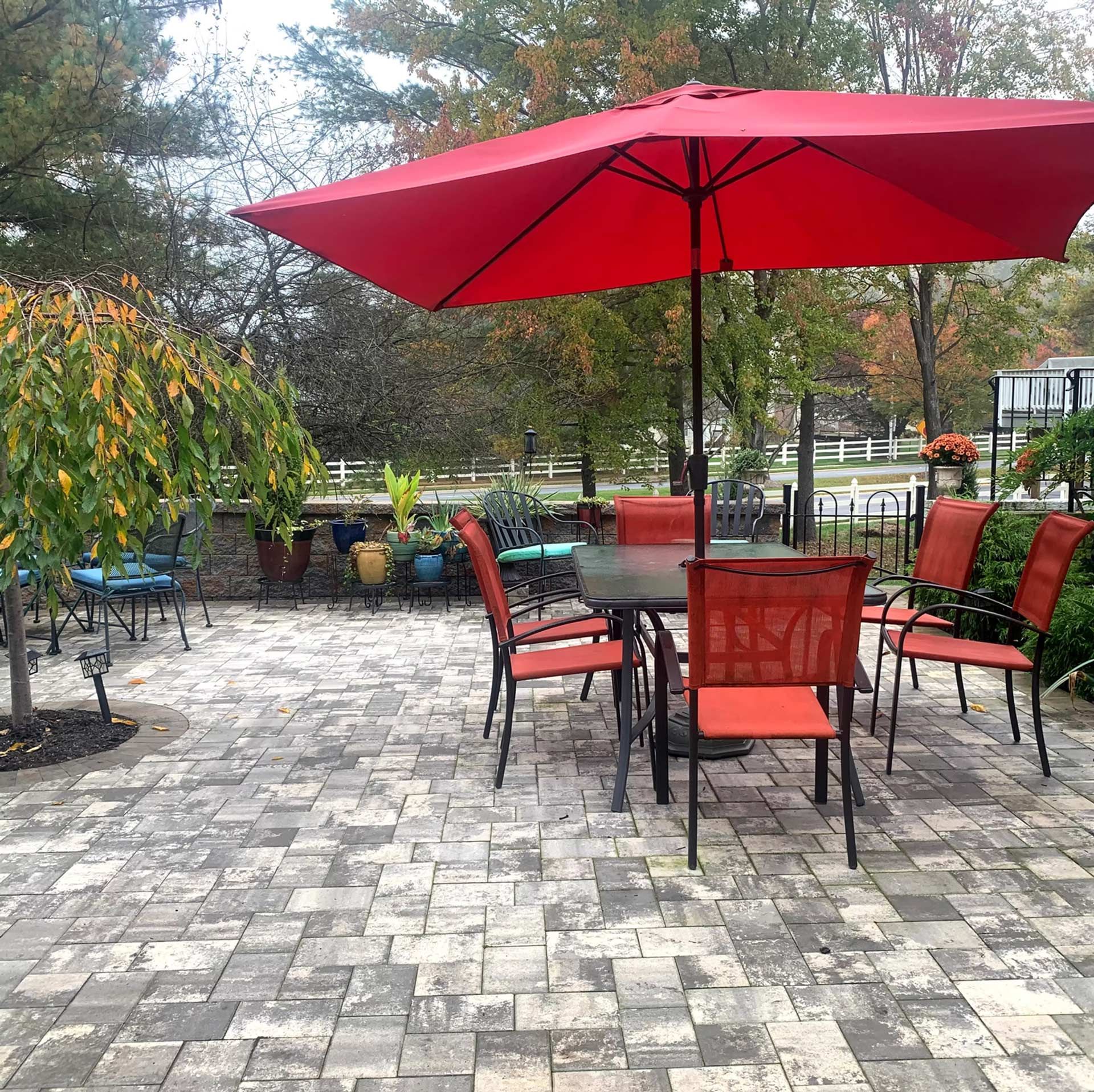A patio set with red chairs and a matching umbrella, situated on a stone-paved terrace surrounded by trees and greenery.