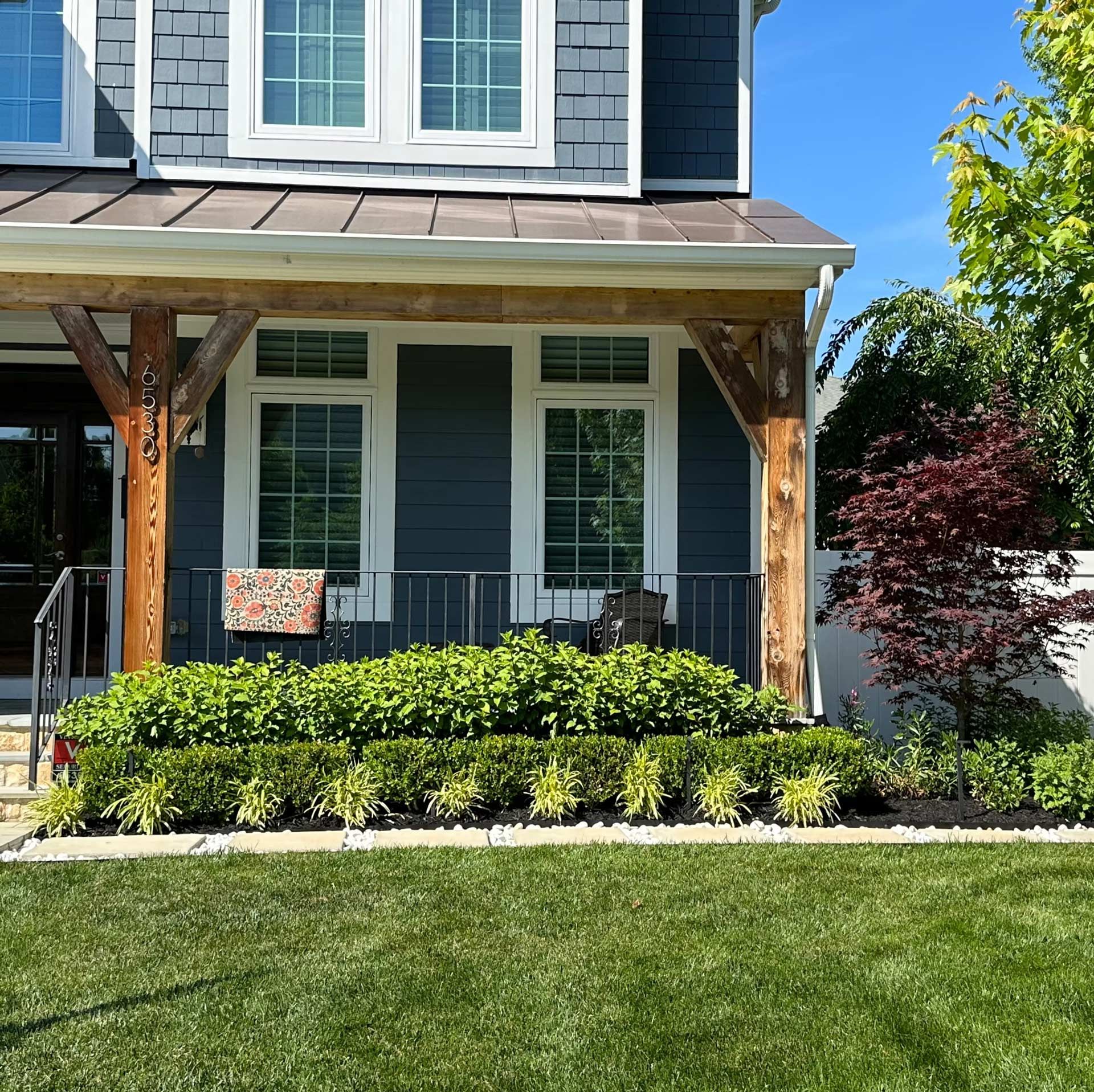 A blue house with a wooden porch, white-trimmed windows, and a foundation garden featuring green shrubs and small trees.