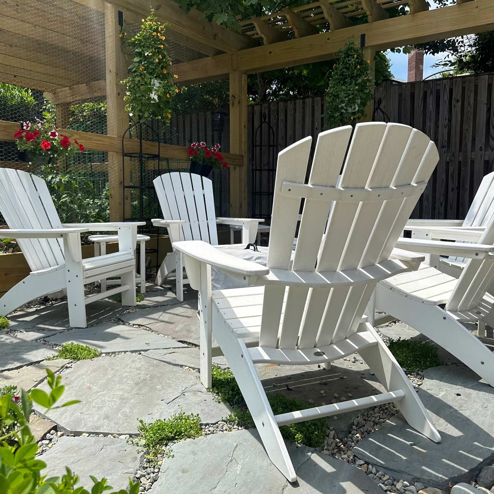 White Adirondack chairs arranged on a stone patio beneath a wooden pergola in a garden.