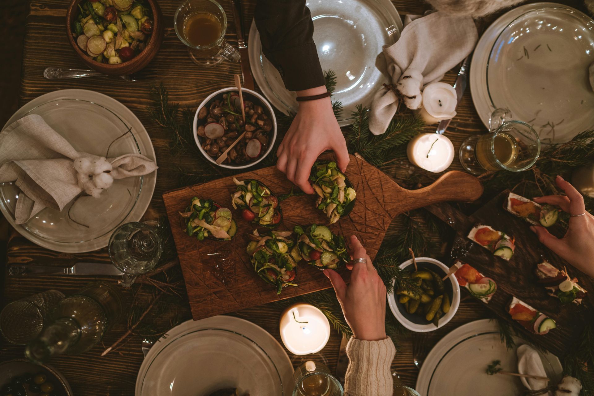 Hands reaching for food on a wooden table set for a meal. Includes plates, bowls, candles, and greenery.