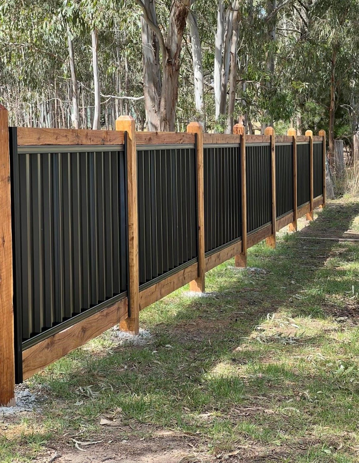 A black fence with wooden posts is surrounded by grass and trees.