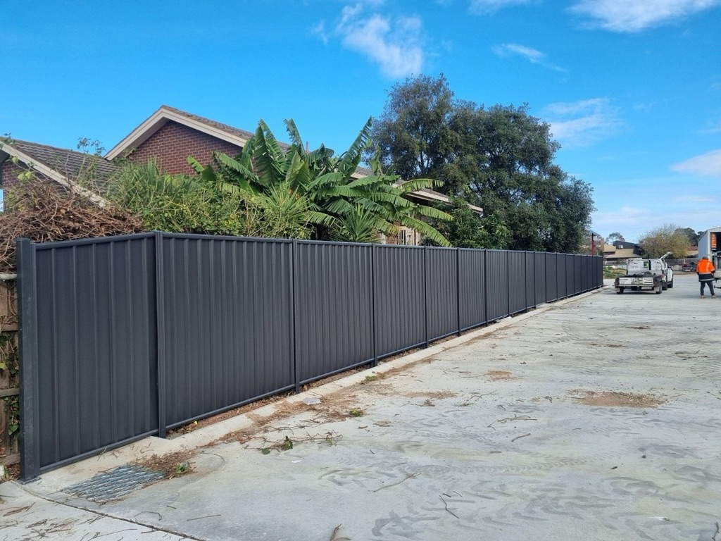 Dark gray corrugated metal fence along a concrete area, trees, blue sky.