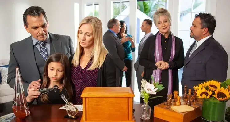 Family at a memorial service: adults, child, wooden urn, and a scattering of guests in a bright room.