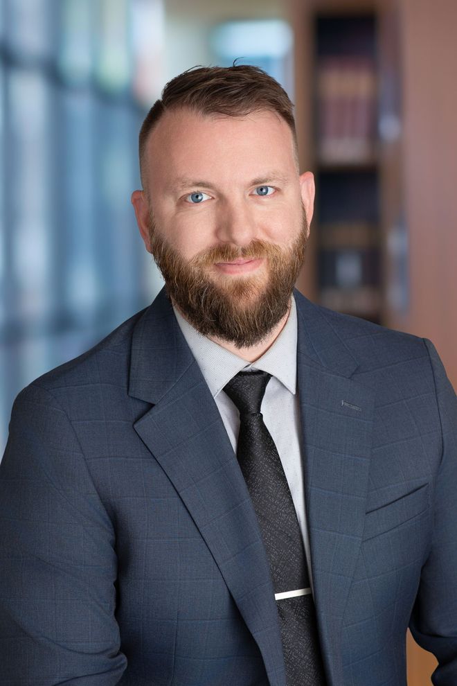 A man with a beard is wearing a suit and tie with his arms crossed.