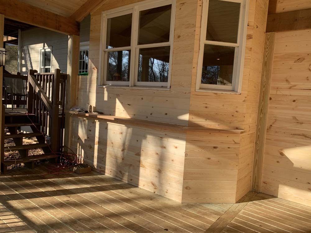 Wooden deck and wall with a window, sunlight casting shadows. A dark wooden staircase is visible on the left.