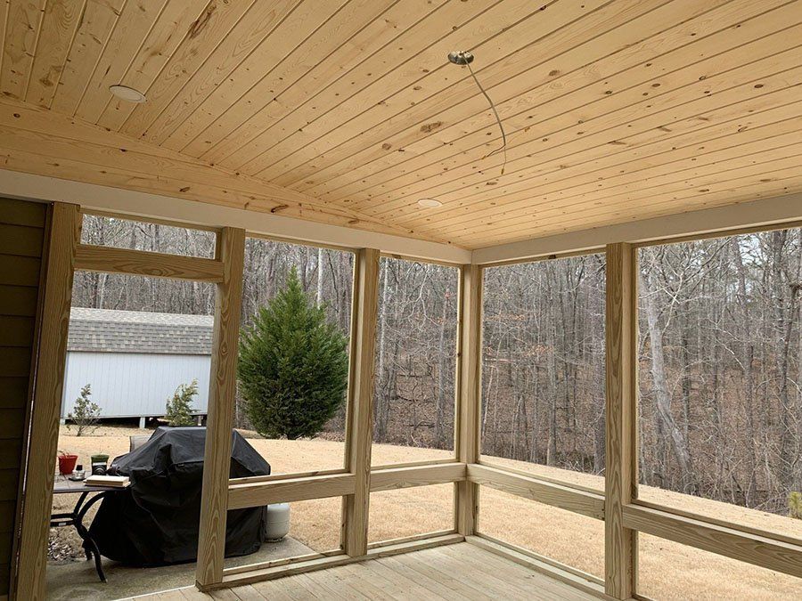 Screened porch interior with a wooden ceiling and framed windows overlooking a wooded area.