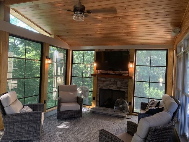 Sunroom with large windows, wood ceiling, fireplace, and wicker furniture. Sunlight streams in, illuminating the space.
