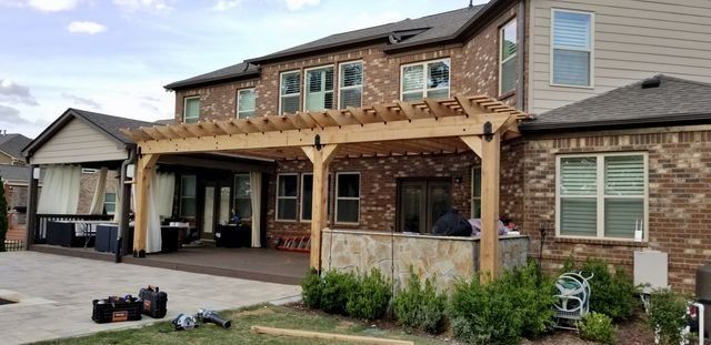 A backyard patio with a wooden pergola is attached to a multi-story brick house. Shrubs line the base of the patio.