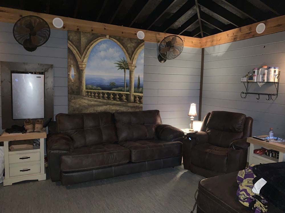A cozy living space with brown leather furniture, a large mural, and a small refrigerator. Two fans hang from the ceiling.