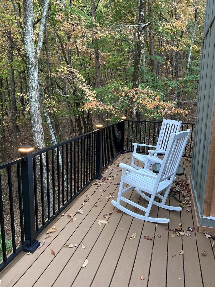 Two white rocking chairs sit on a wooden deck overlooking a forest. The railing is black with lights on top.