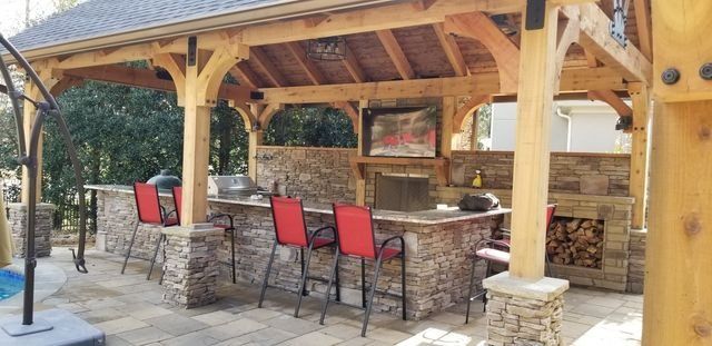 An outdoor kitchen with a stone facade. It has a bar with red chairs, a grill, a fireplace, and a wooden roof.