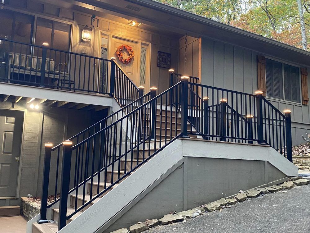 House with black railing and lit posts, with a wreath on the door and fall foliage in the background.