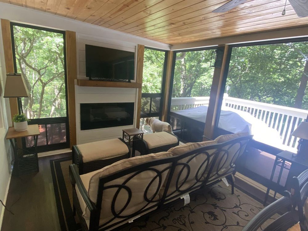 A screened porch with a sofa, fireplace, and TV. Large windows offer a view of lush green trees and a deck.