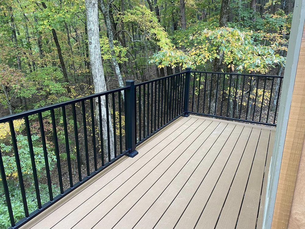 A wooden deck with black railings overlooks a forest. Light brown deck boards contrast with the dark railing and green trees.