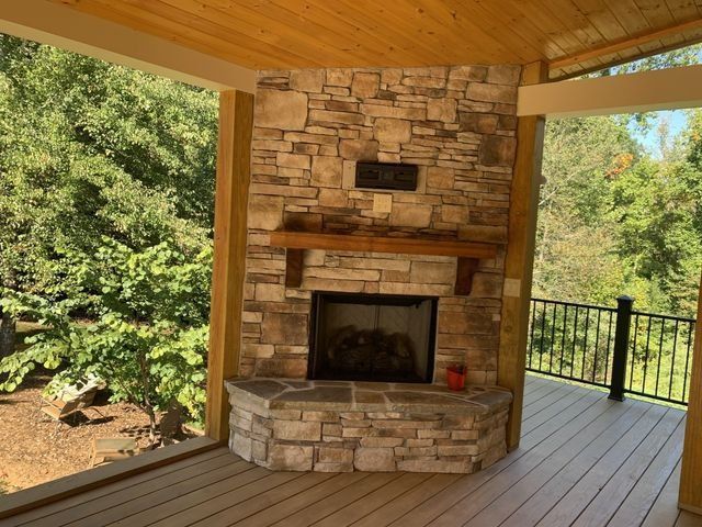 Stone fireplace on a wooden deck, surrounded by trees. The fireplace has a wooden mantel and a dark hearth.