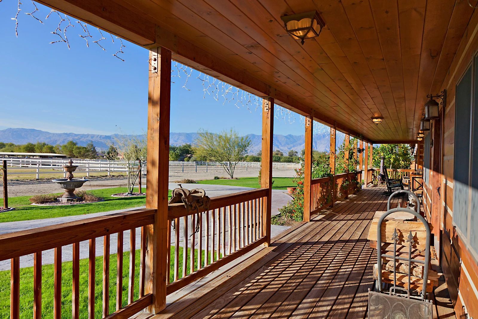 Wooden porch of a home with a view of a green field, fountain, and distant mountains under a blue sky.