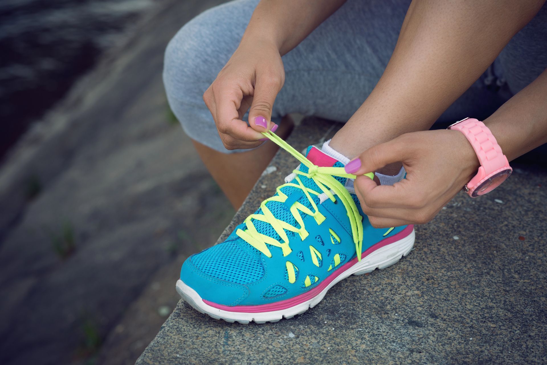 Mujer atando una zapatilla deportiva de color azul brillante y rosa con cordones amarillos en un escalón de hormigón.
