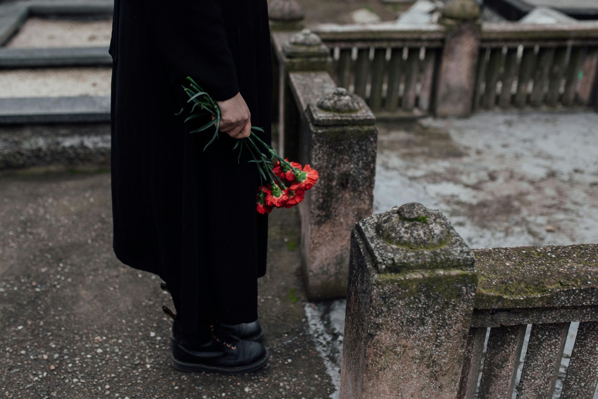 A man in a black coat is holding a bouquet of red roses in a cemetery.