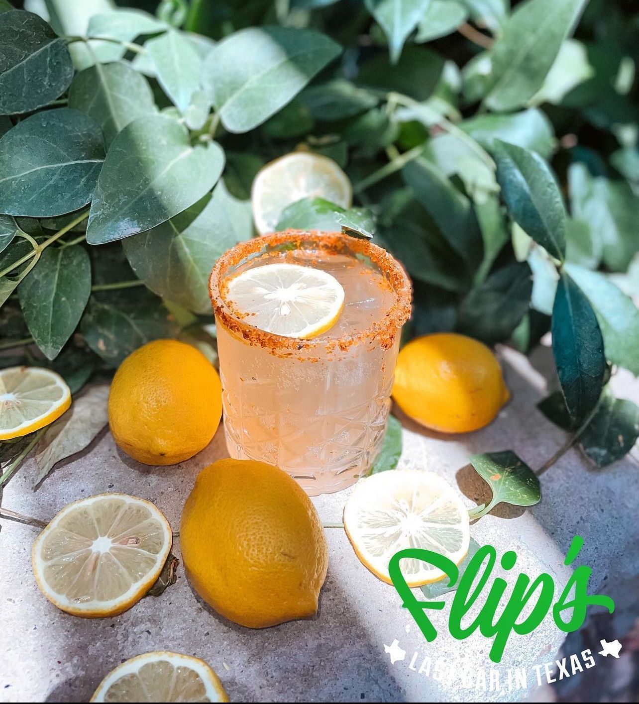 A drink with a slice of lemon in it is surrounded by lemons and leaves.