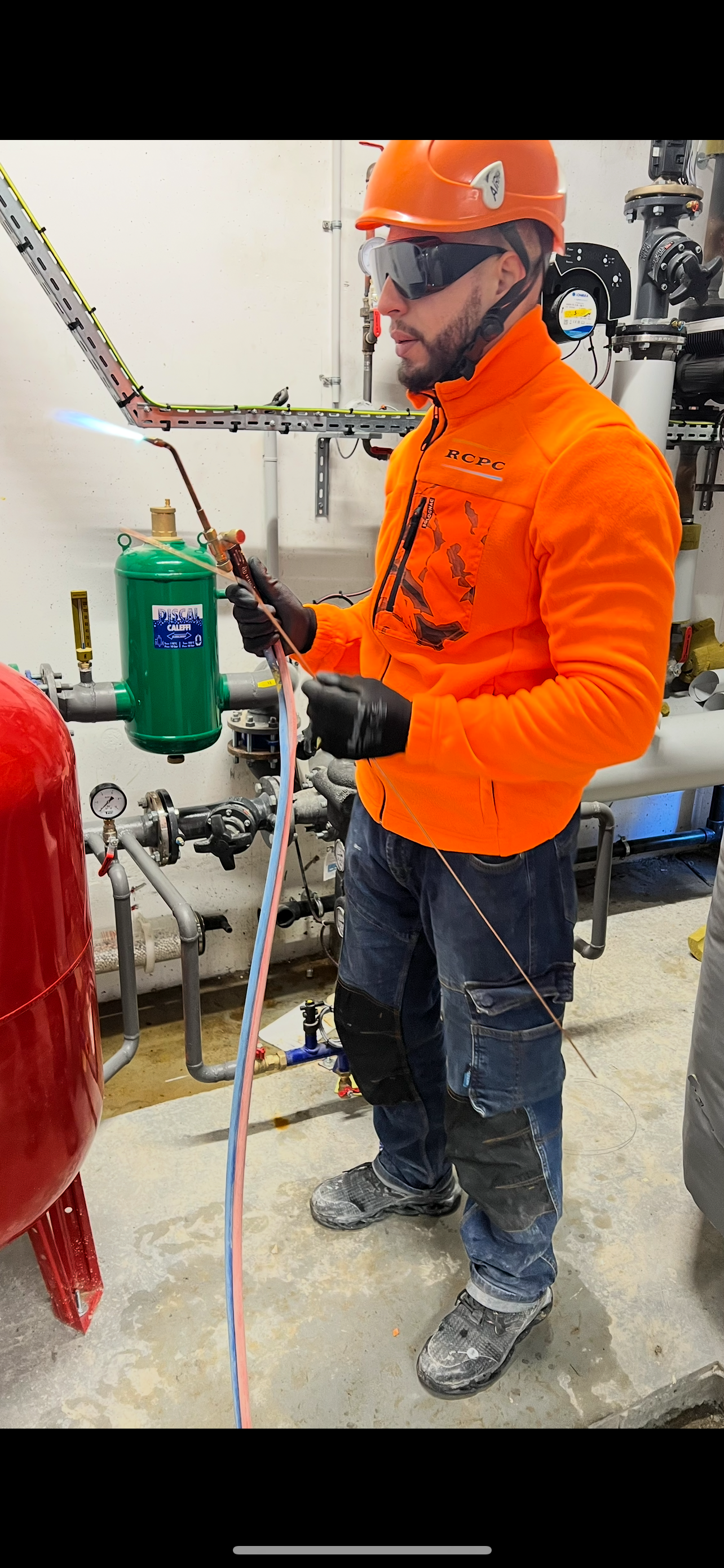 Un ouvrier en tenue de sécurité orange soude des tuyaux avec un chalumeau, portant des lunettes de sécurité et un casque de sécurité, en intérieur.
