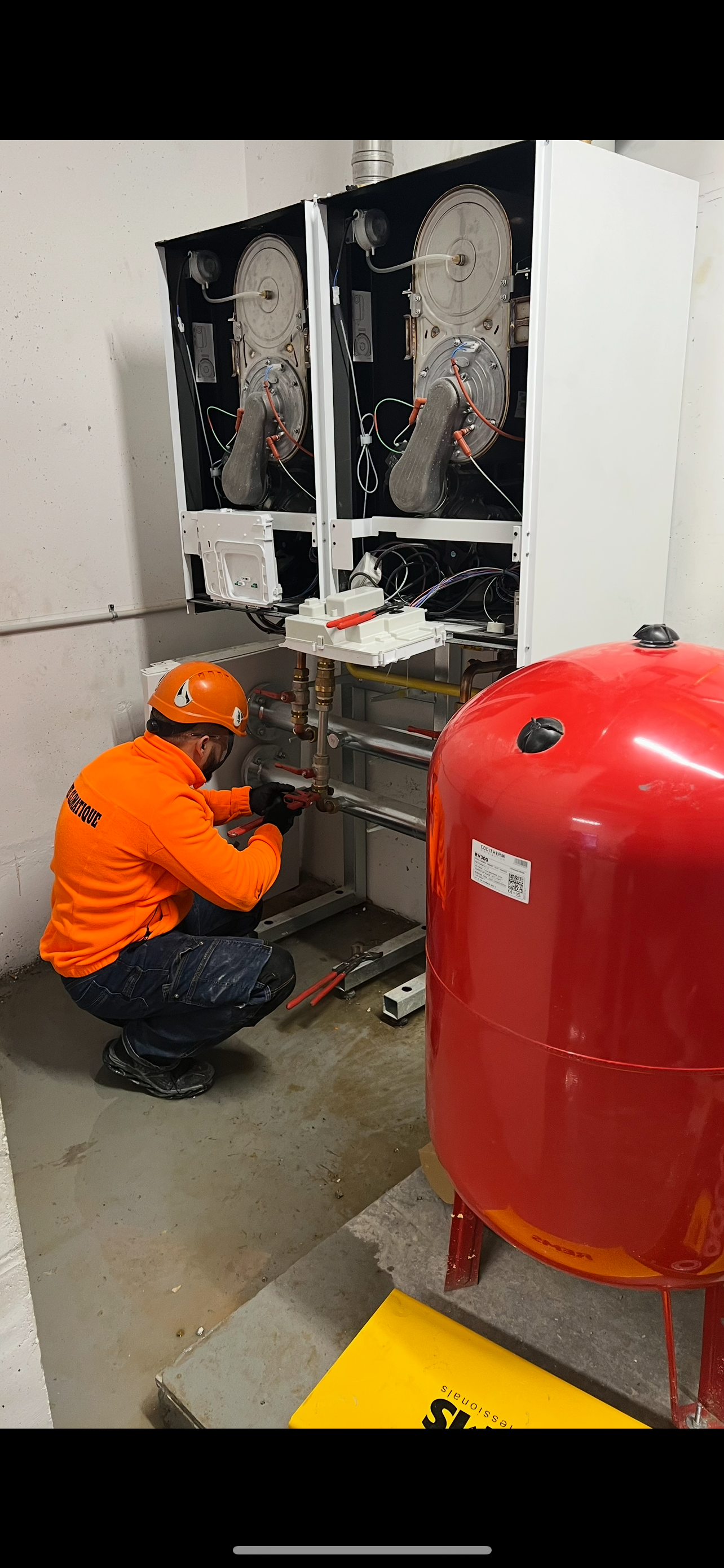 Un ouvrier en salopette orange est accroupi près d'un réservoir rouge et de machines blanches, à l'intérieur d'un bâtiment.