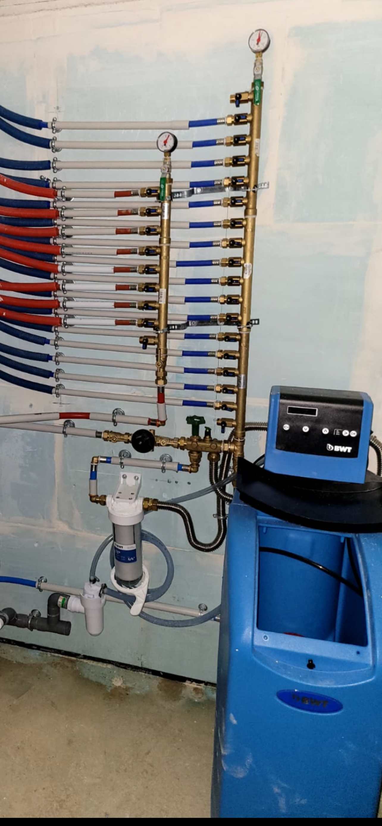 Système de plomberie complexe avec tuyaux, vannes et manomètres montés sur un mur, reliés à un adoucisseur d'eau bleu.