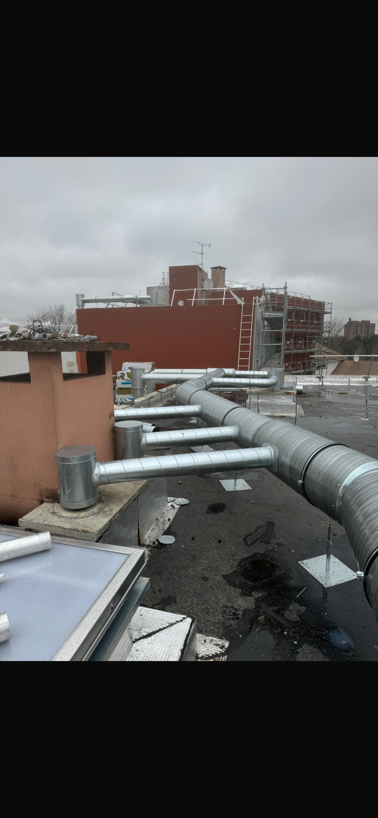 Toit avec des tuyaux de ventilation argentés et des bâtiments en arrière-plan sous un ciel couvert.