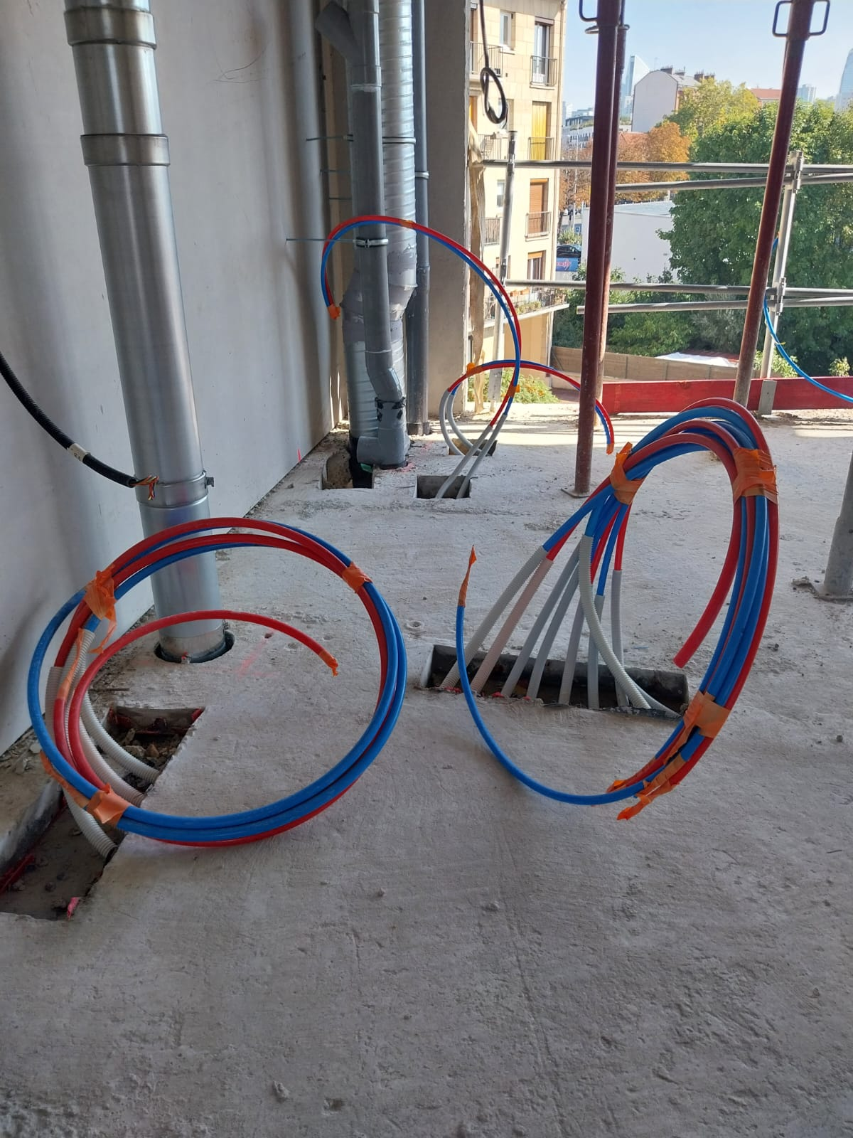 Des tuyaux spiralés rouges et bleus émergent des ouvertures du sol en béton. Environnement de chantier, près de tuyaux métalliques et d'échafaudages.