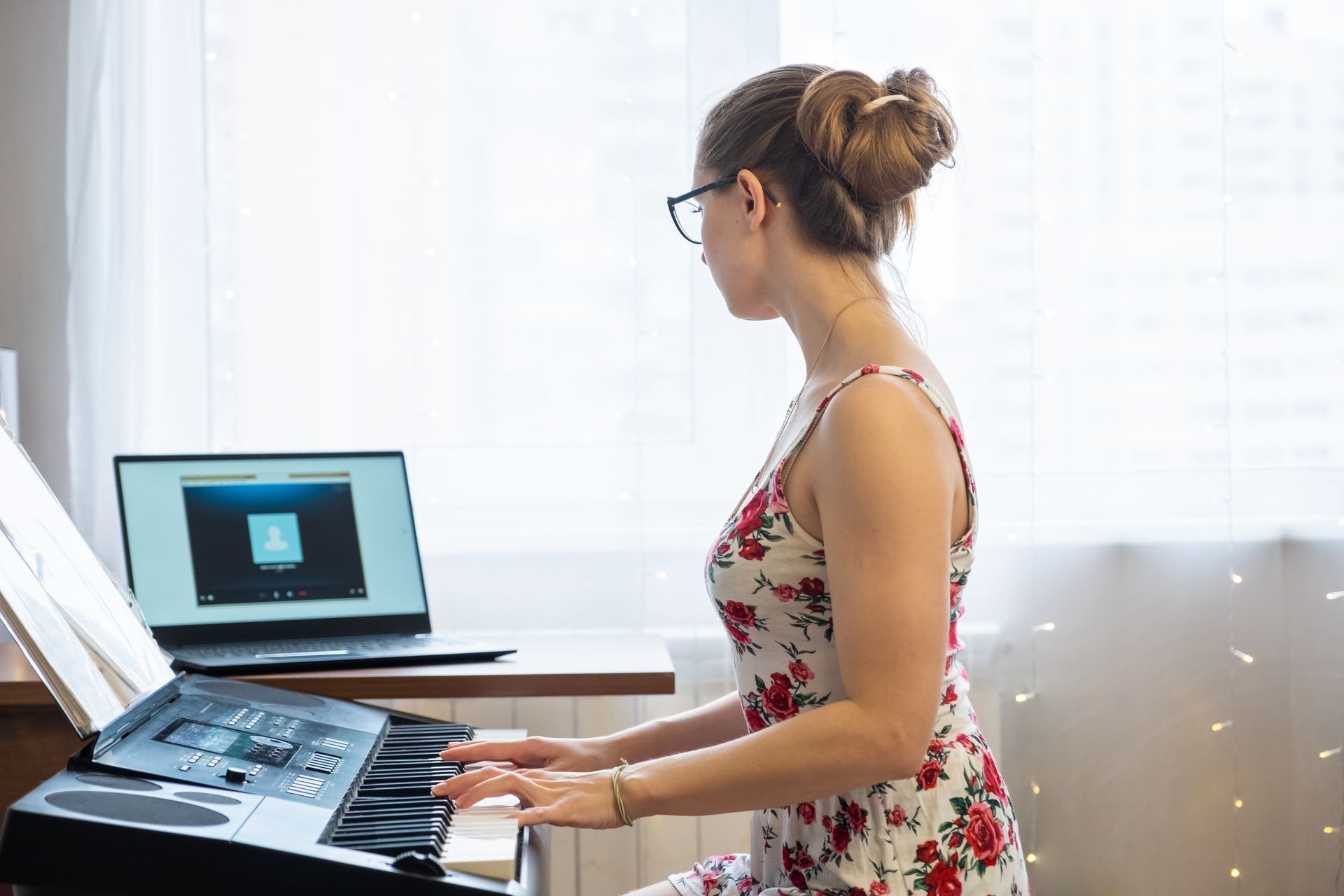 Woman playing a keyboard with laptop open on a desk by a window. She wears glasses and a floral dress.