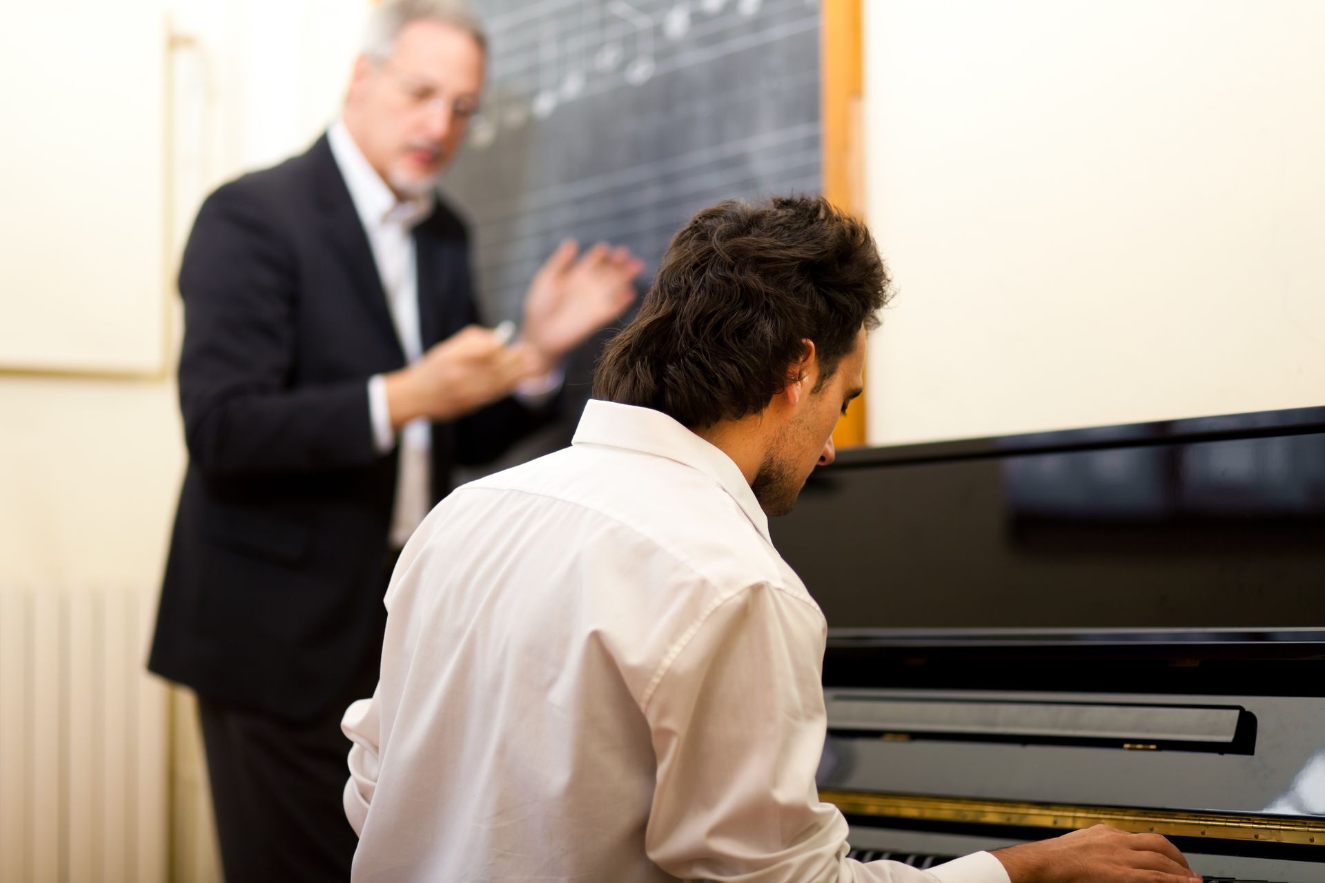 A piano student with a teacher, who gestures behind the student, in a music classroom.