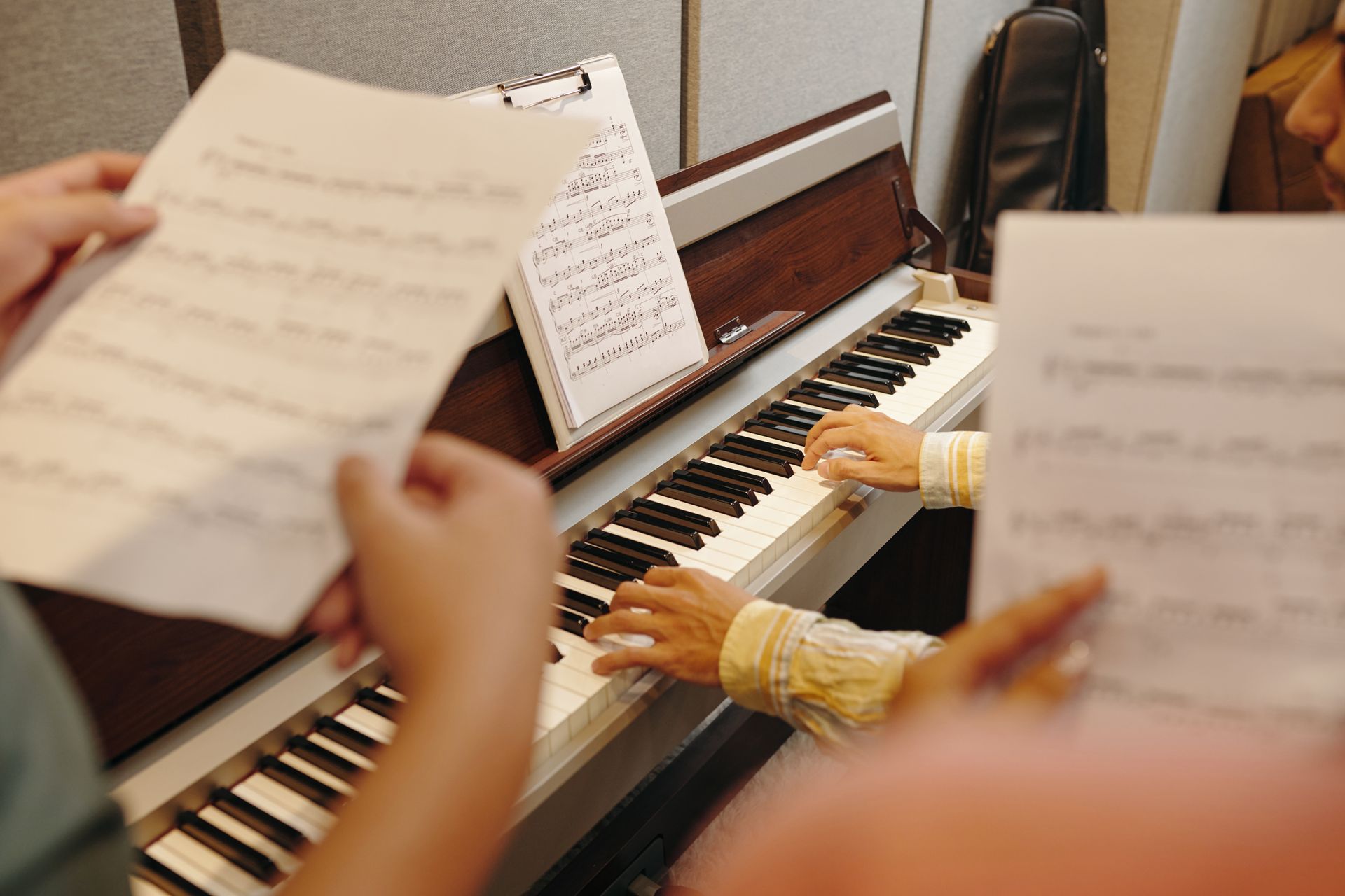 Person playing a piano with sheet music; two others hold sheet music nearby in a recording studio.