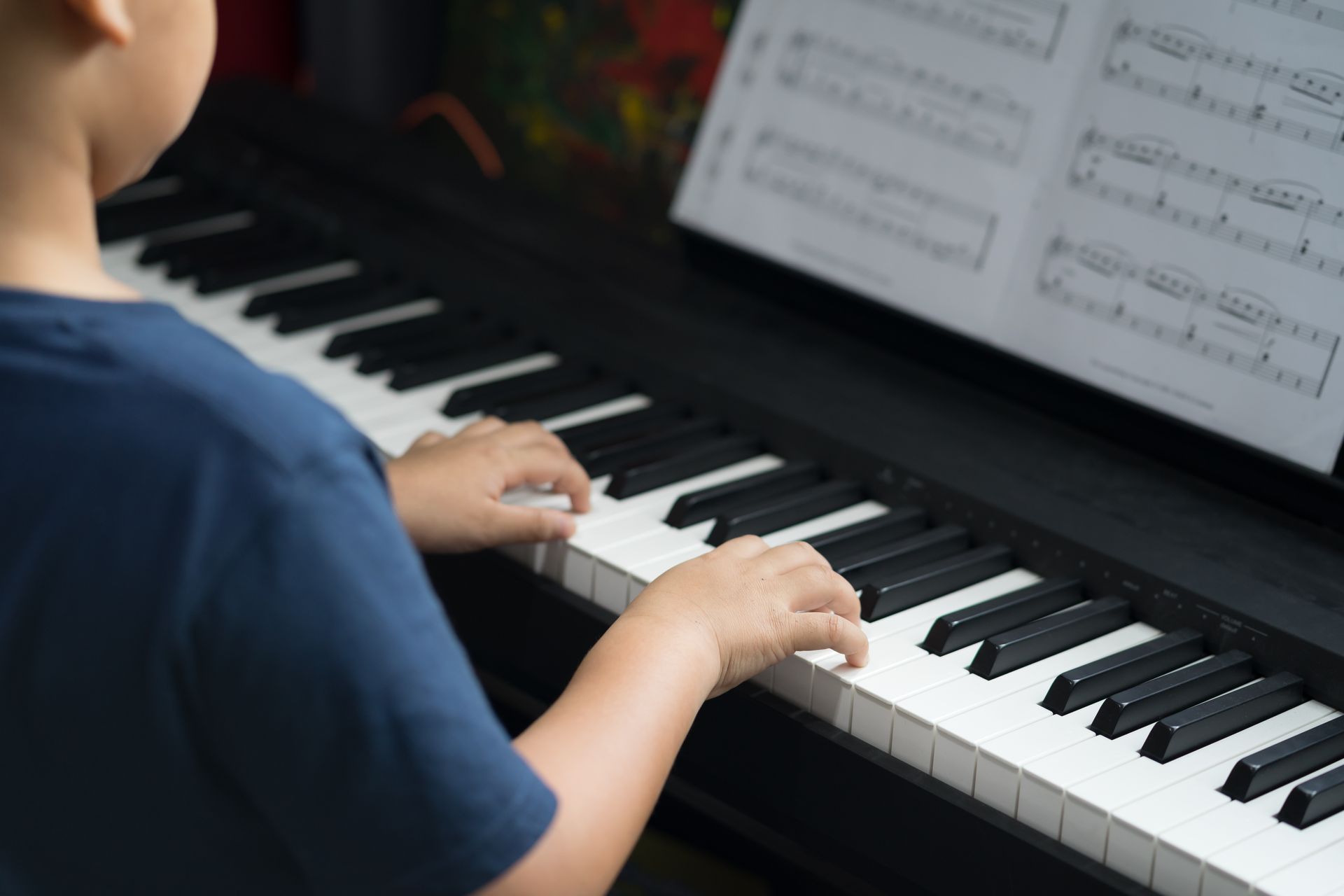 A child's hands playing a black piano, following sheet music. They wear a blue shirt in a room.