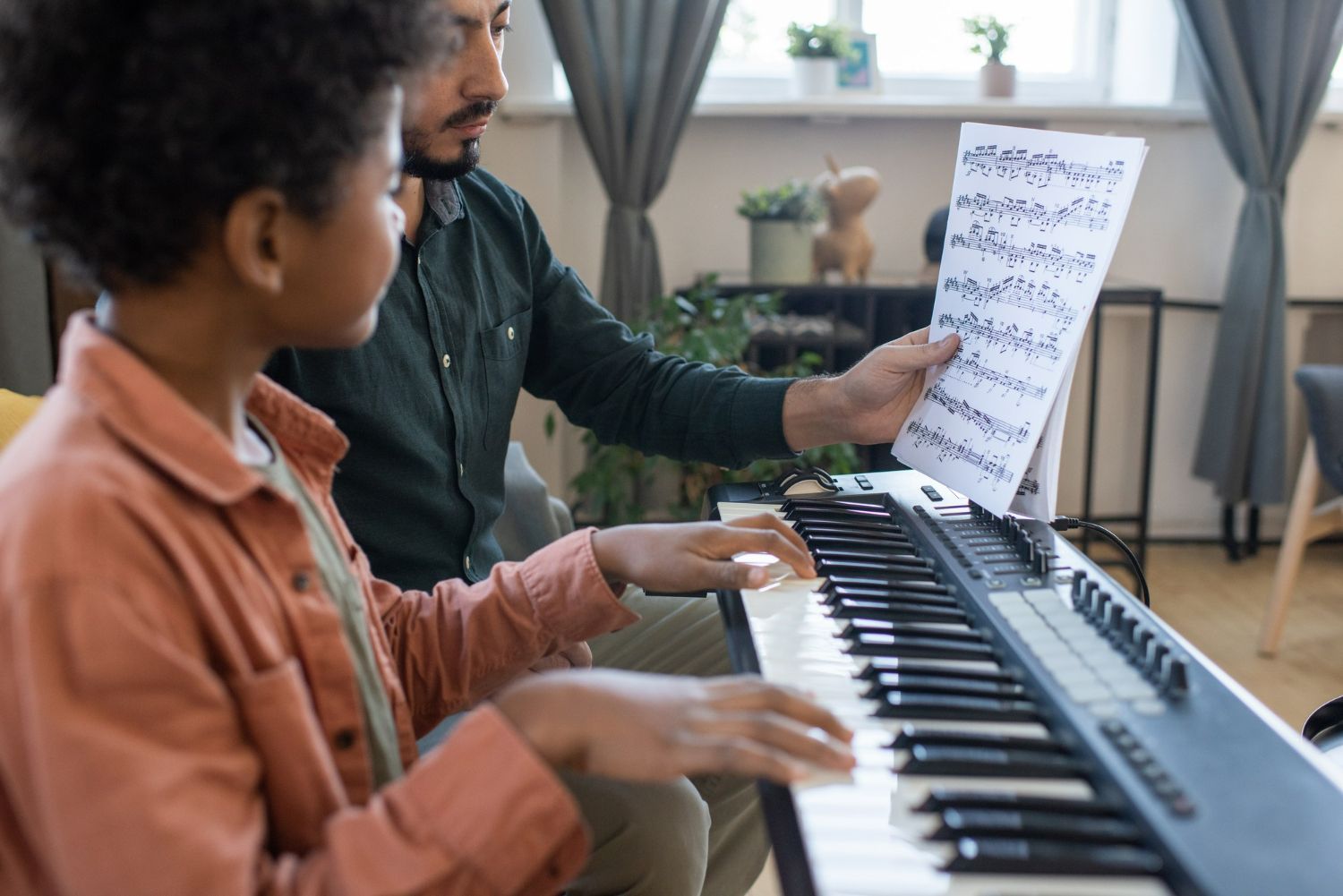 Man teaching a Black child to play piano, pointing at sheet music indoors.