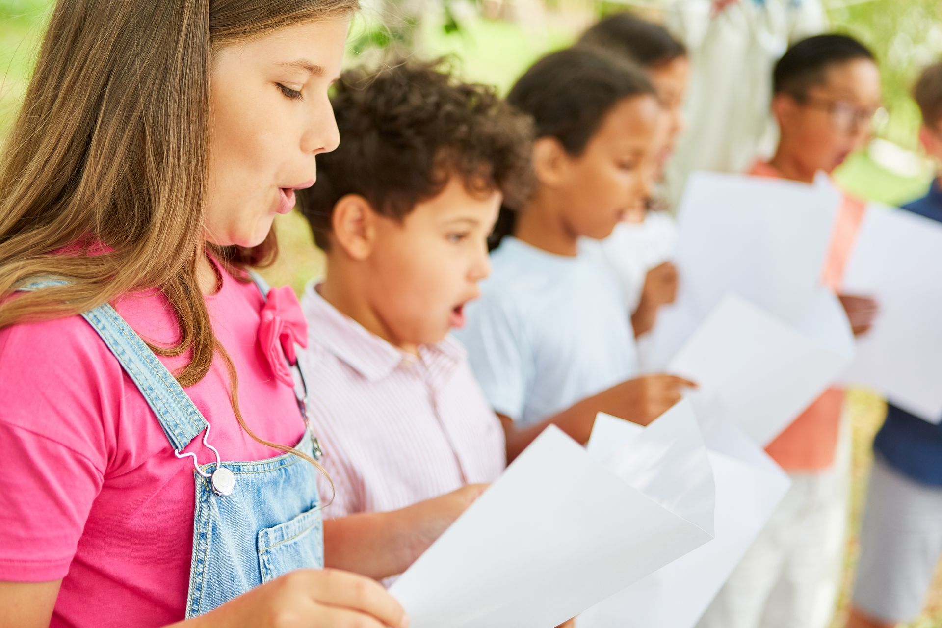 Children in a row outdoors, reading white sheets of paper during a group activity