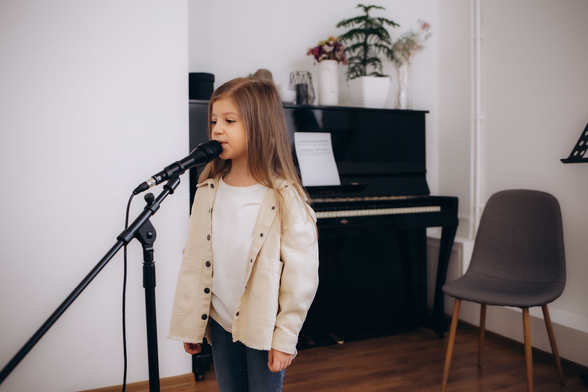 Girl singing into microphone near a piano. She wears a beige jacket and stands in a bright room.