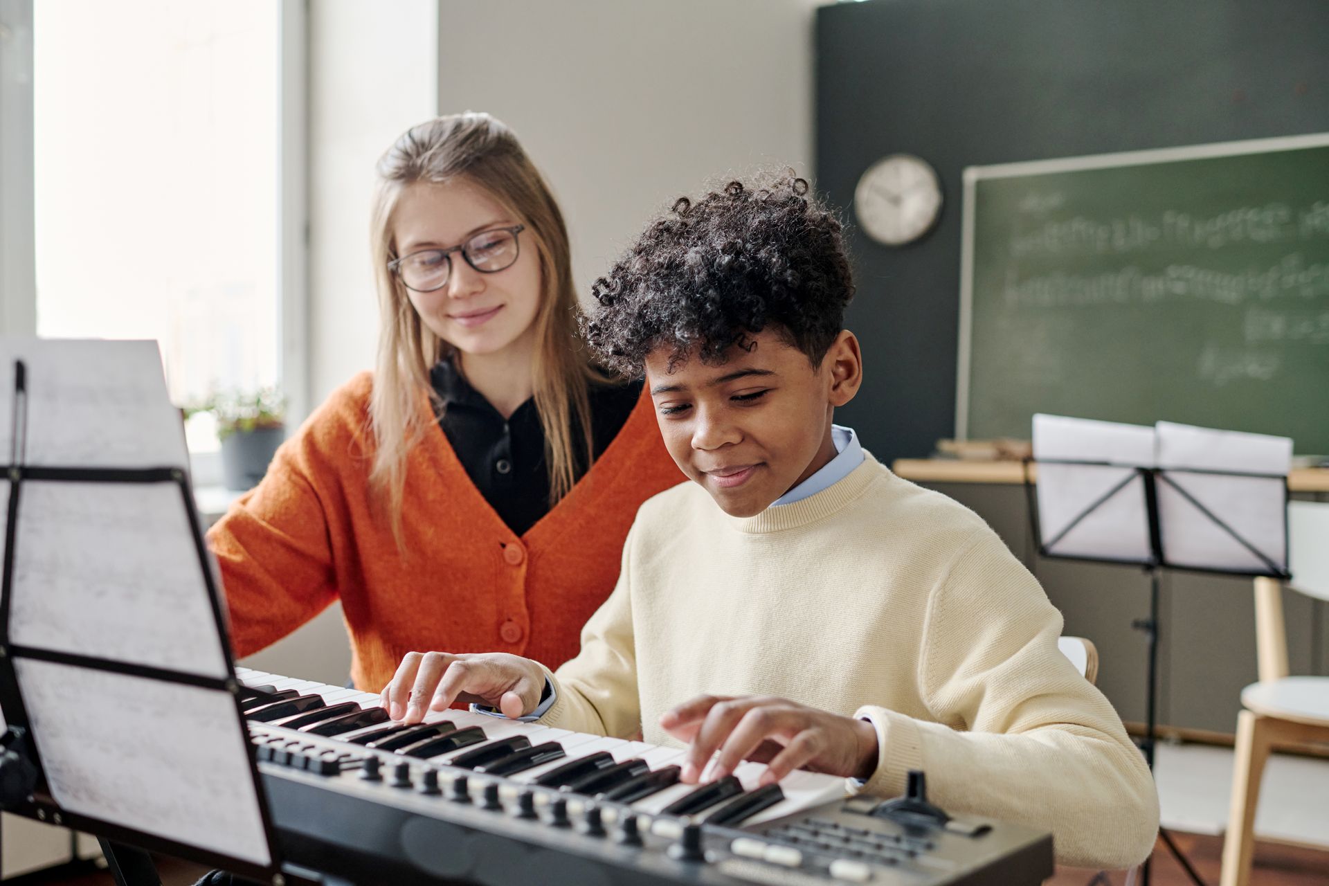 Two people playing a keyboard in a classroom, with sheet music stands and a chalkboard behind them.