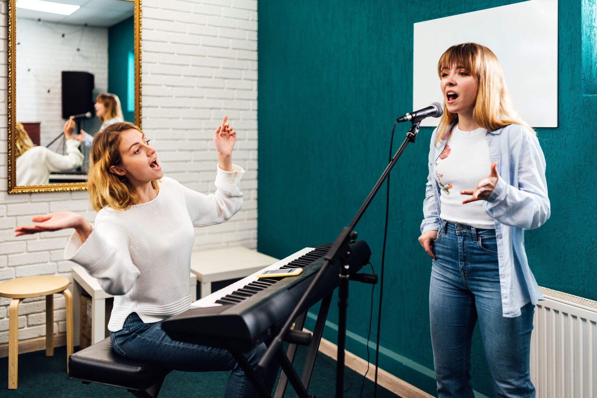 Two young women in a recording studio; one singing into a microphone, the other at a keyboard gesturing.