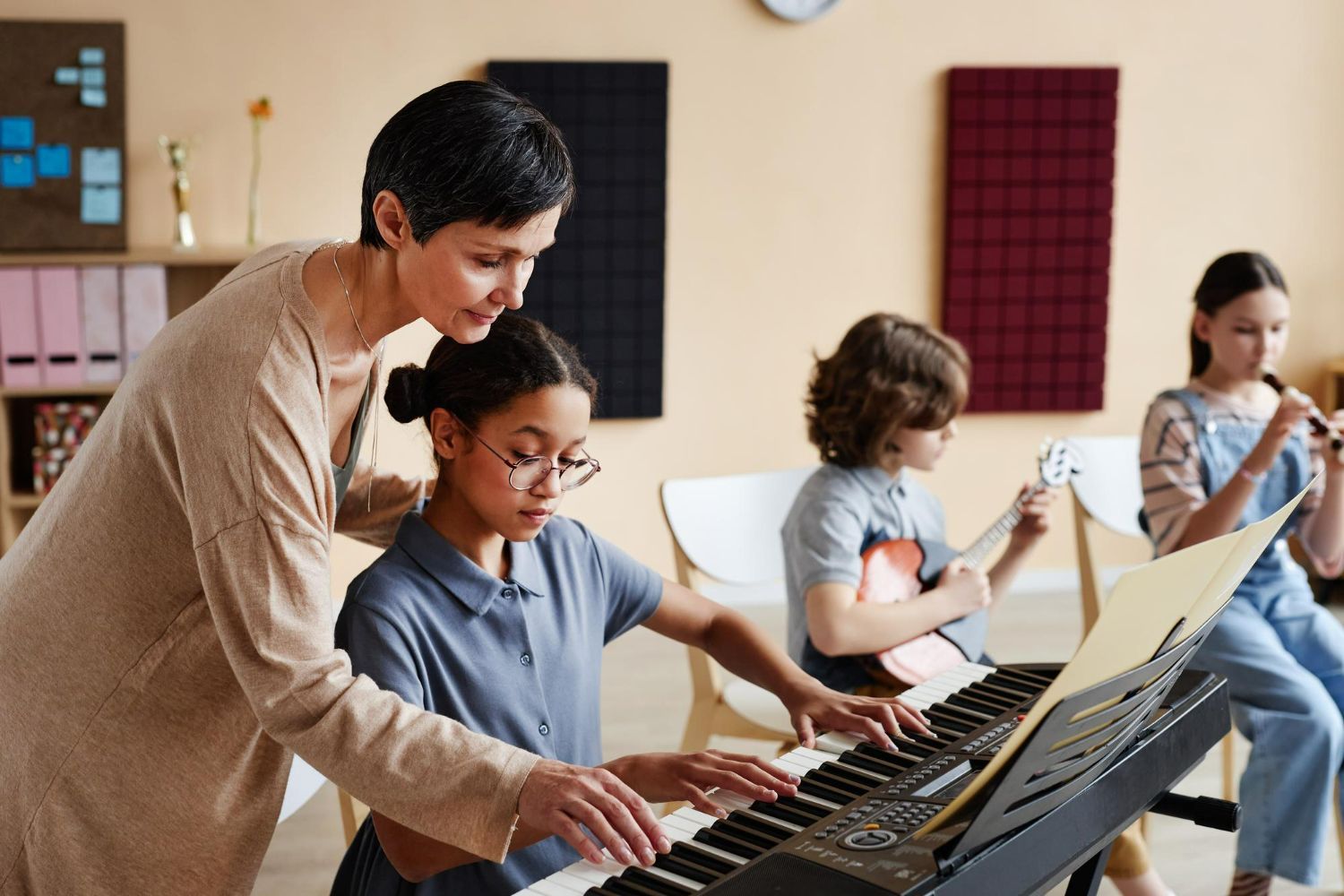 Teacher helping student play keyboard in music class. Other students play guitar and flute.