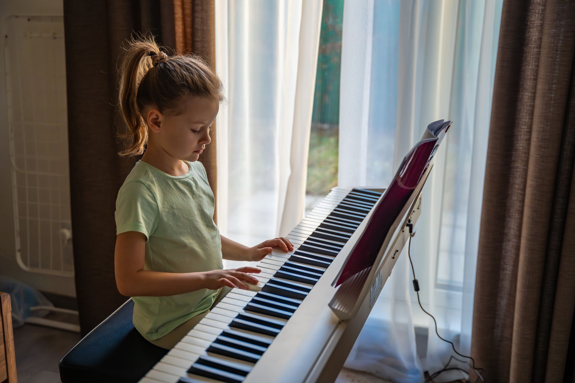 Child playing a digital piano by a sunlit window, wearing a light green shirt.