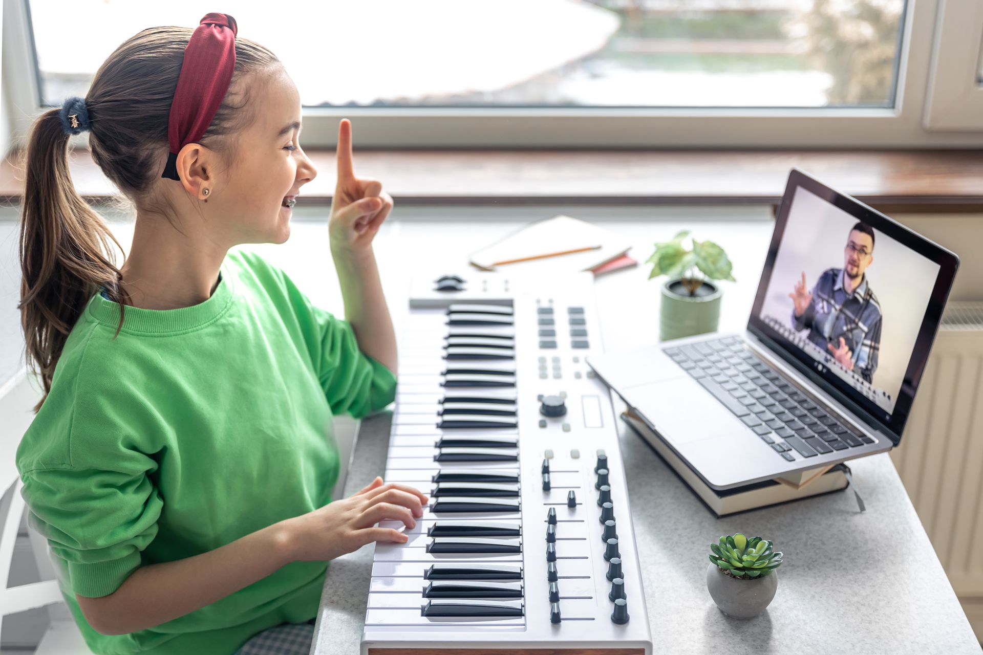 A young girl learns piano online with a laptop, pointing upwards. She wears a green shirt and red headband in a bright room.