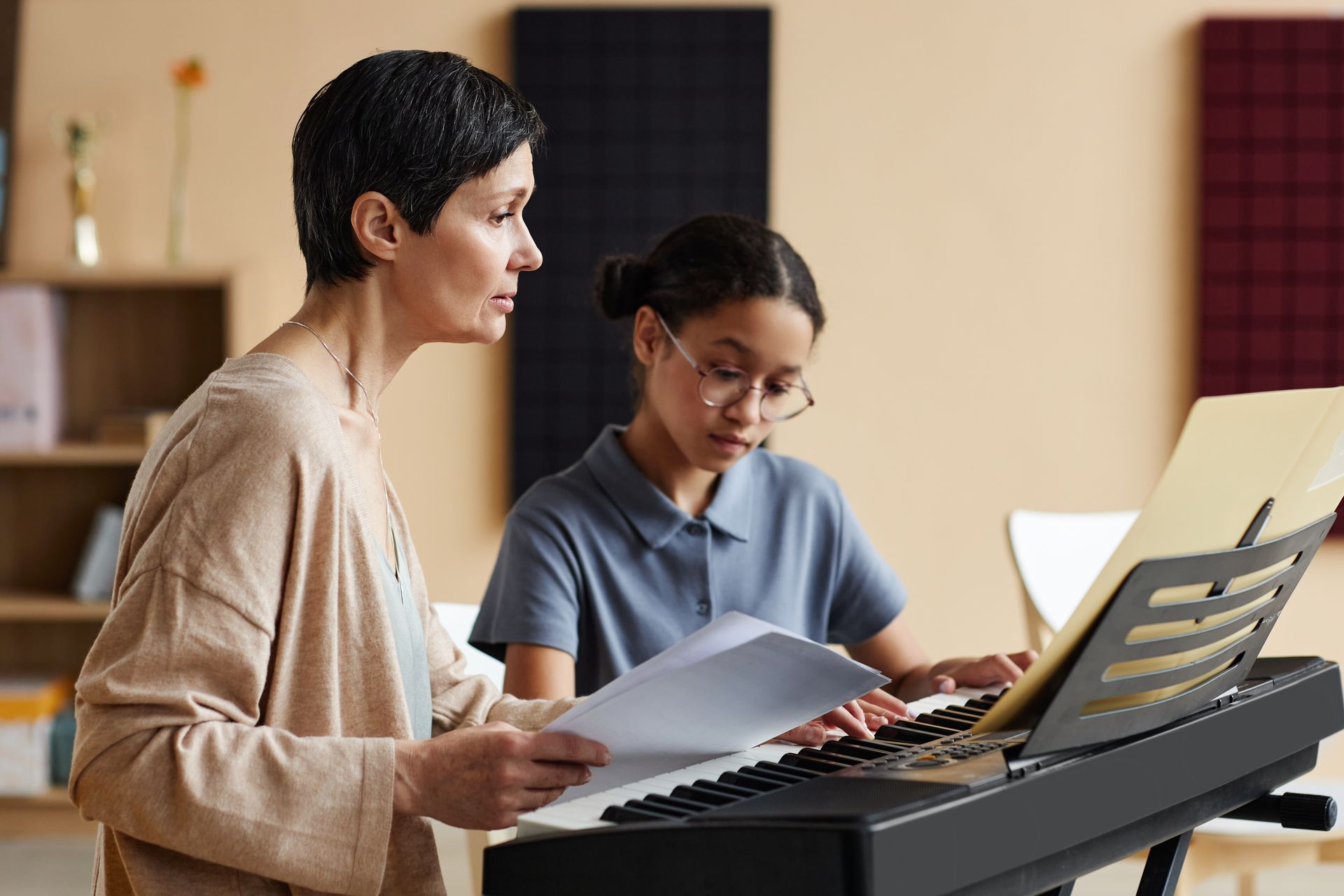 A woman teaches a girl piano. They're in a music room, with the girl playing while wearing glasses.