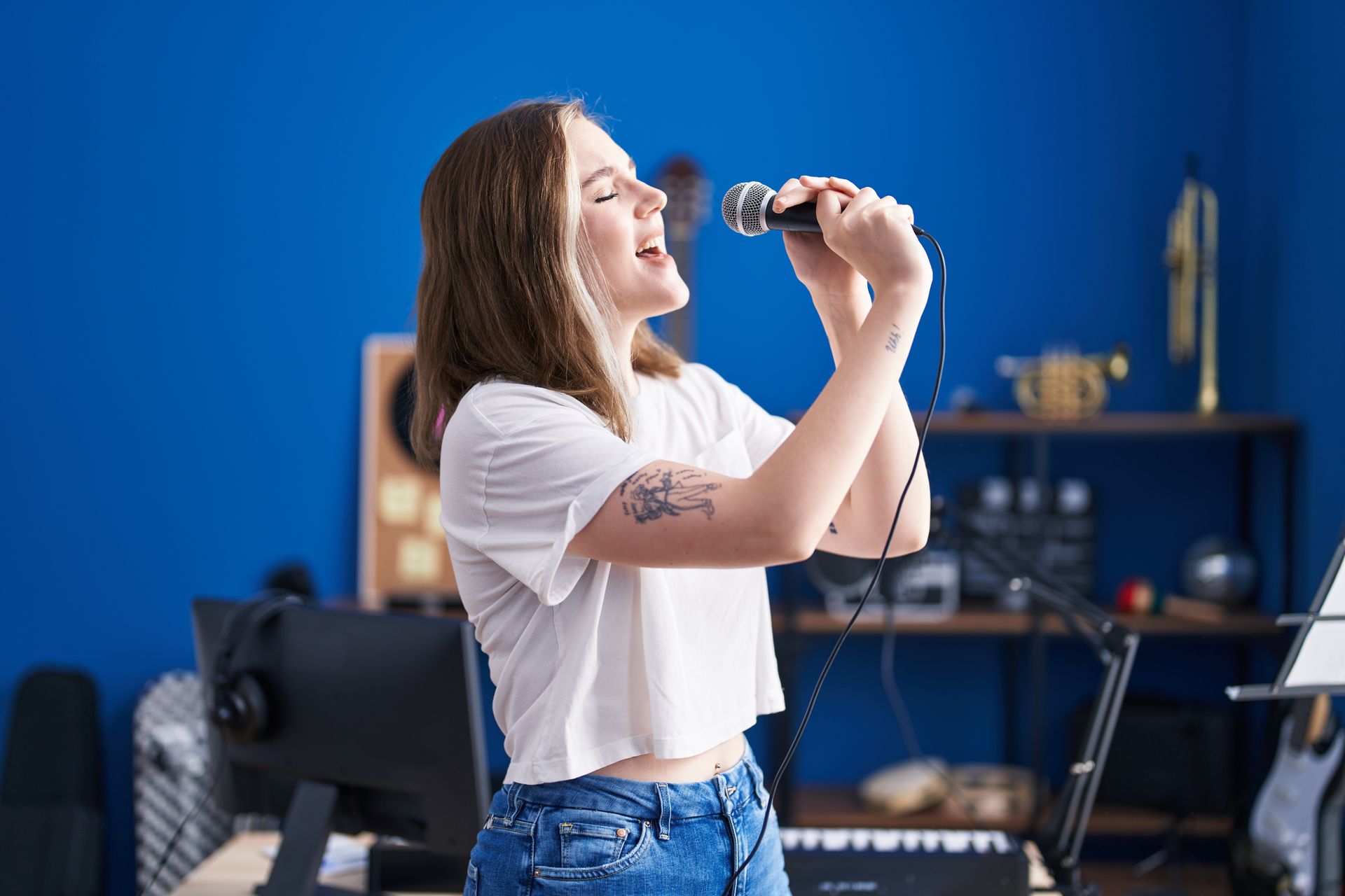 Young woman singing into a microphone in a music studio. She wears a white shirt and jeans, with a tattoo on her arm. The room has a blue wall.
