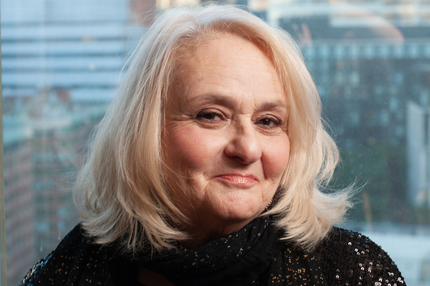An individual with light hair and a soft smile, wearing a dark, shimmering top against a blurred city background.
