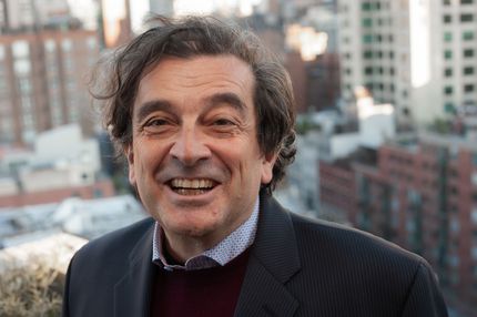 A smiling man with curly dark hair wearing a dark blazer over a collared shirt, with a city building background.