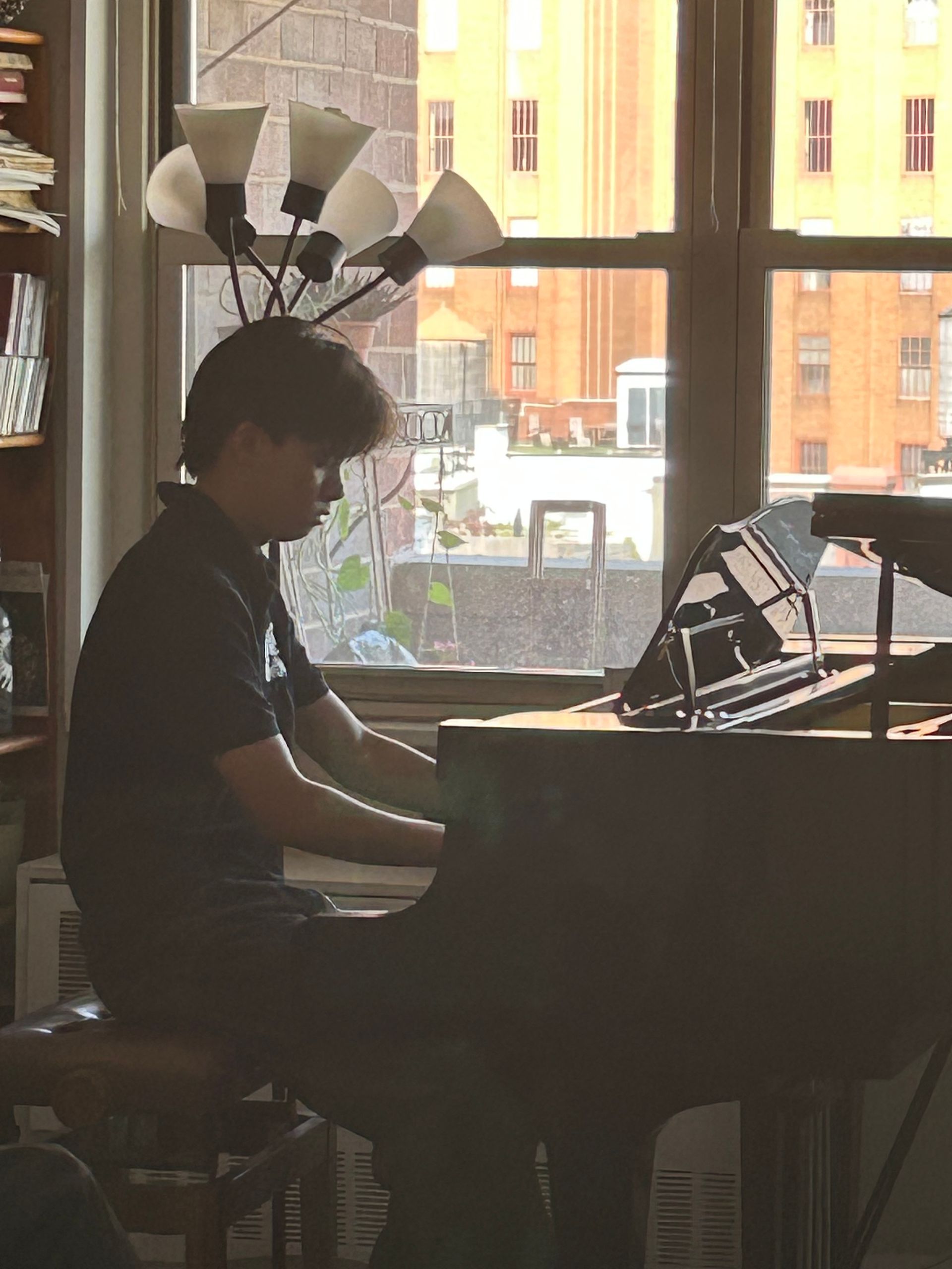 Person playing a grand piano in a dim room by a large window.